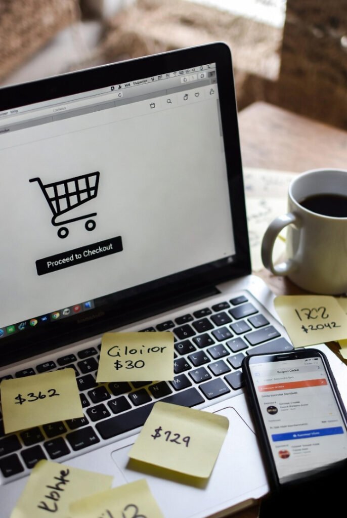 laptop open to a checkout page, phone with coupon tabs, coffee mug, and sticky notes with scribbled codes.